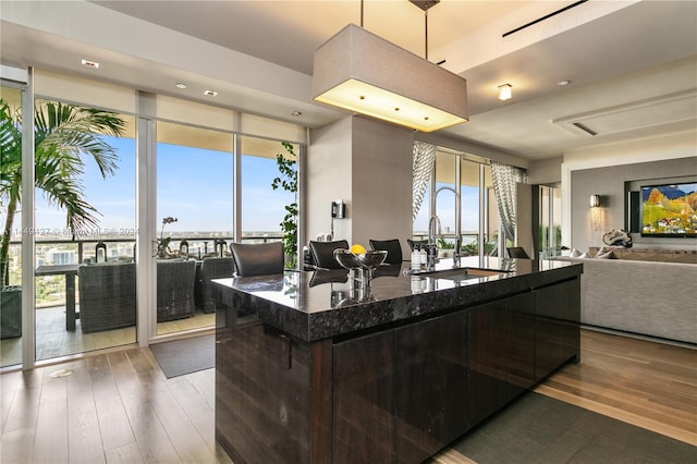 kitchen with an island with sink, sink, dark stone counters, floor to ceiling windows, and hardwood / wood-style floors