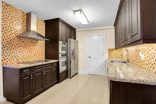 kitchen featuring wall chimney range hood, light stone countertops, appliances with stainless steel finishes, dark brown cabinetry, and sink