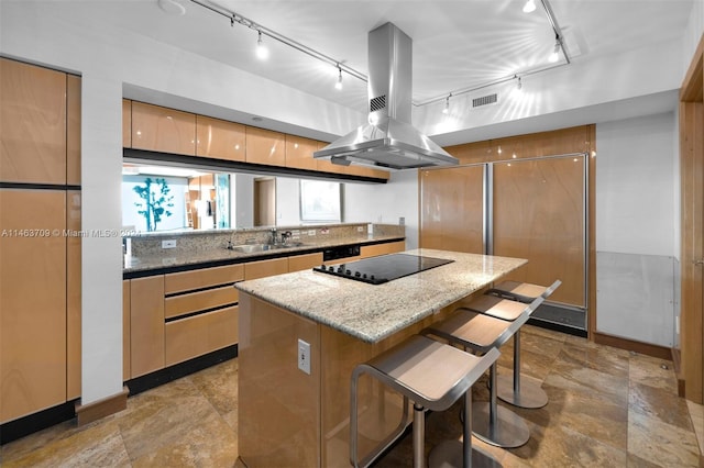 kitchen featuring sink, island exhaust hood, light stone counters, a breakfast bar area, and black electric cooktop