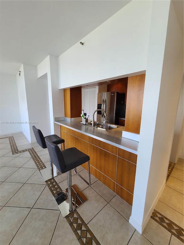 kitchen featuring light tile flooring, stainless steel counters, a breakfast bar area, stainless steel fridge, and sink