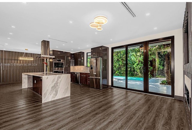 kitchen featuring a breakfast bar, island range hood, an island with sink, stainless steel appliances, and dark wood-type flooring