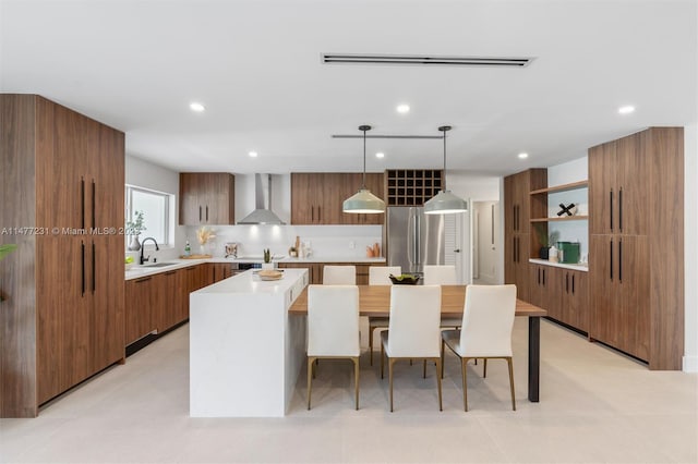 kitchen with wall chimney range hood, hanging light fixtures, sink, a center island, and stainless steel refrigerator