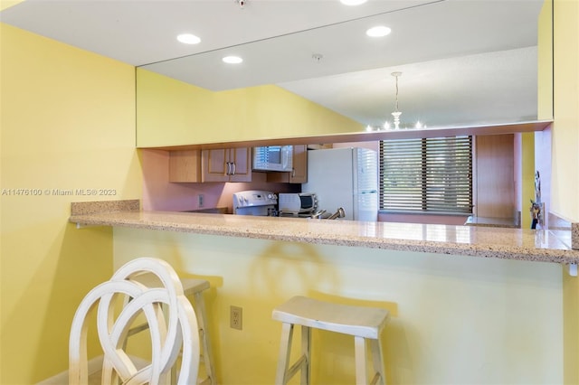 kitchen featuring kitchen peninsula, white appliances, a chandelier, light stone countertops, and a kitchen bar