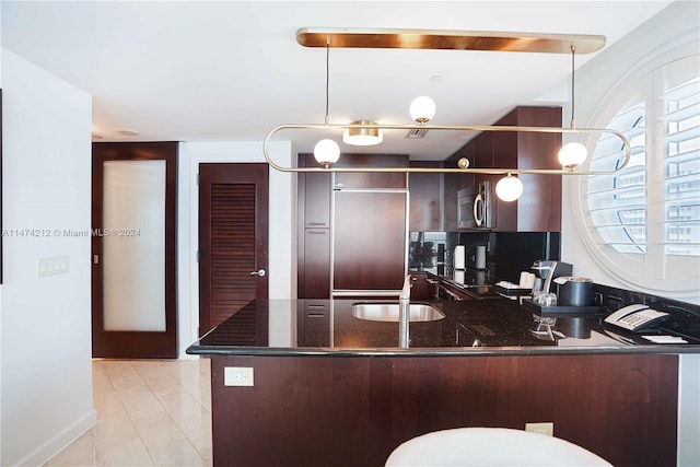 kitchen featuring light tile flooring, kitchen peninsula, dark brown cabinets, hanging light fixtures, and dark stone countertops