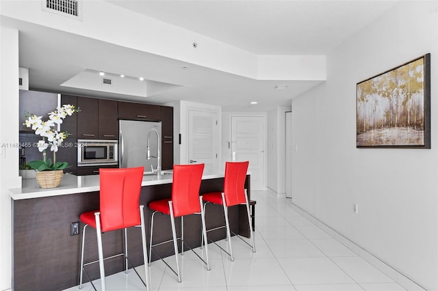 kitchen with stainless steel appliances, light tile flooring, a breakfast bar area, dark brown cabinetry, and a raised ceiling