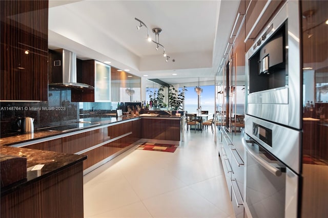 kitchen with backsplash, light tile floors, dark stone counters, rail lighting, and wall chimney exhaust hood