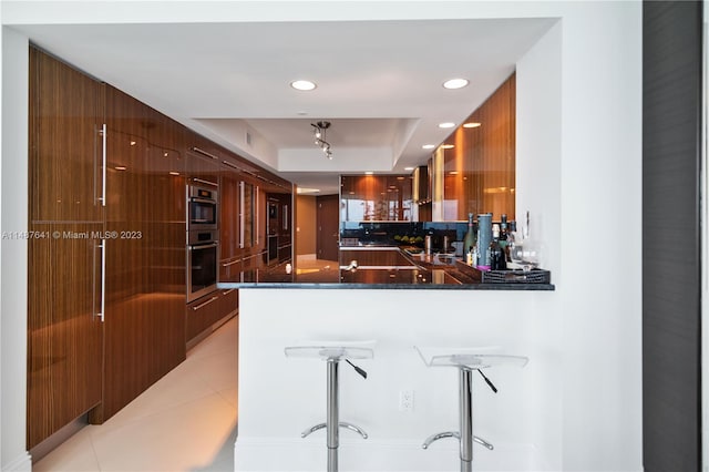 kitchen featuring light tile flooring, dark stone counters, kitchen peninsula, multiple ovens, and a raised ceiling