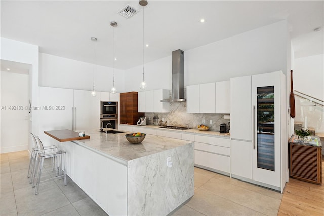 kitchen featuring decorative light fixtures, wall chimney exhaust hood, white cabinetry, backsplash, and a kitchen island with sink