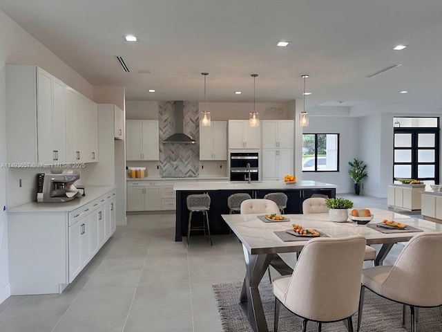 kitchen with white cabinetry, a kitchen breakfast bar, an island with sink, pendant lighting, and wall chimney range hood