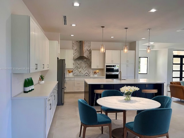 kitchen with an island with sink, wall chimney range hood, freestanding refrigerator, double oven, and light countertops