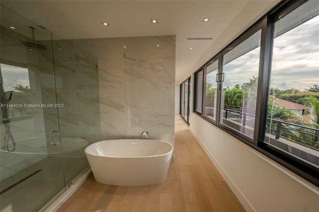 bathroom featuring wood finished floors, a soaking tub, a marble finish shower, and tile walls