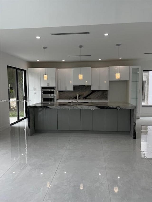 kitchen with double oven, tasteful backsplash, white cabinets, dark stone counters, and hanging light fixtures