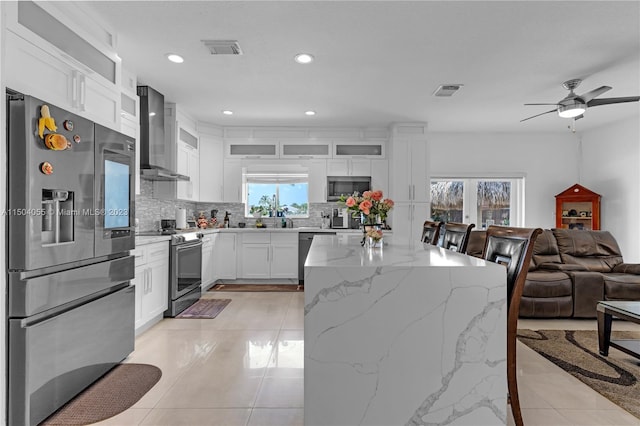 kitchen with wall chimney range hood, stainless steel appliances, backsplash, ceiling fan, and white cabinetry