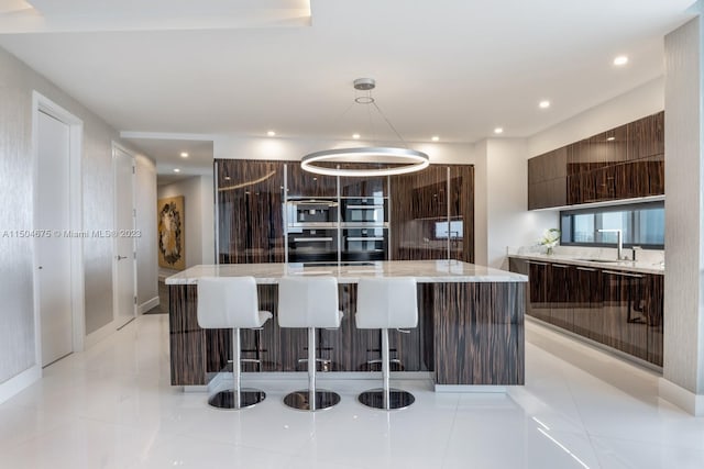 kitchen with an island with sink, stainless steel double oven, pendant lighting, and light tile floors