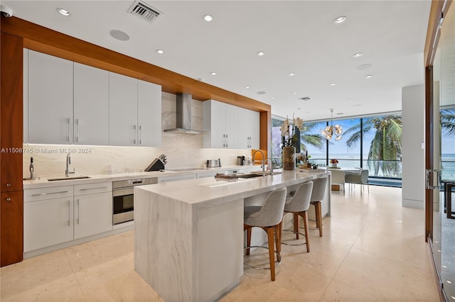 kitchen featuring oven, wall chimney exhaust hood, a kitchen island with sink, and white cabinets