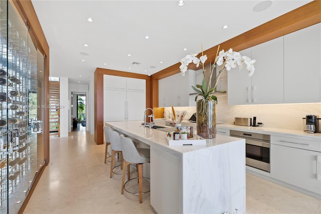 kitchen with white cabinetry, sink, stainless steel oven, decorative backsplash, and a center island with sink