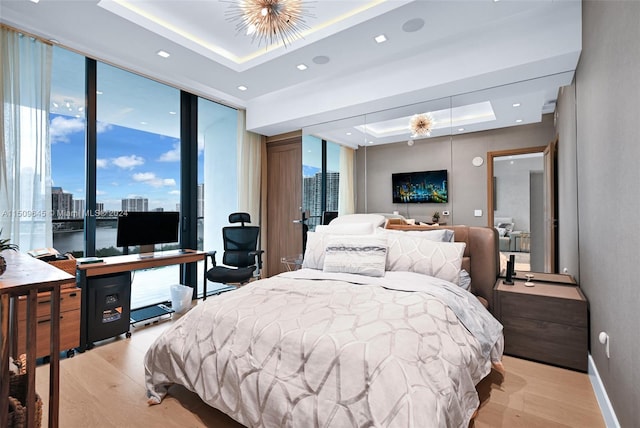 bedroom featuring light wood-type flooring, an inviting chandelier, a water view, expansive windows, and a raised ceiling