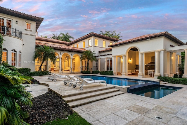 back house at dusk featuring a patio area, a swimming pool with hot tub, and a balcony