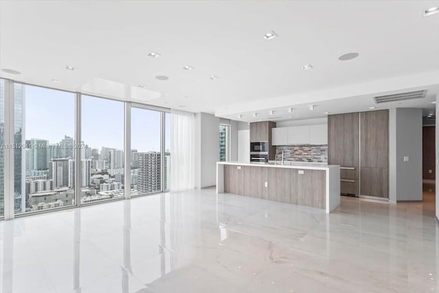 kitchen featuring an island with sink, a wall of windows, white cabinets, light tile flooring, and tasteful backsplash