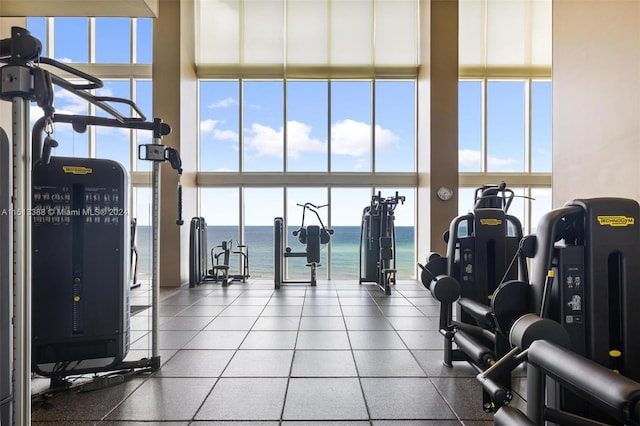 gym featuring a towering ceiling, dark tile flooring, a healthy amount of sunlight, and a water view