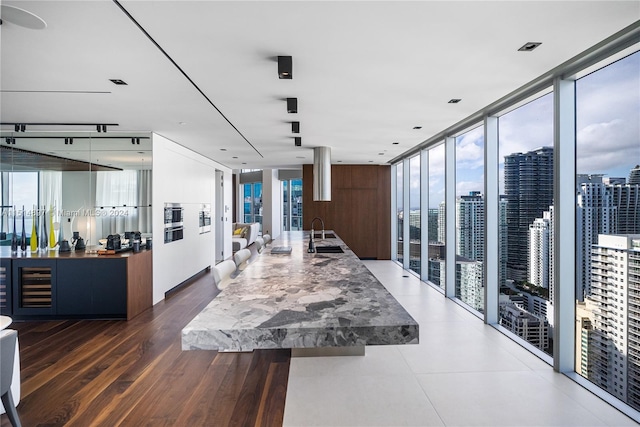 interior space with a wall of windows, a kitchen island with sink, sink, beverage cooler, and light stone counters