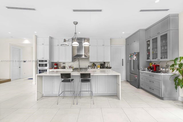 kitchen with light tile flooring, a kitchen island with sink, appliances with stainless steel finishes, a breakfast bar, and wall chimney range hood