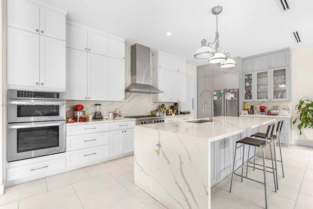 kitchen with an island with sink, hanging light fixtures, appliances with stainless steel finishes, wall chimney range hood, and a notable chandelier