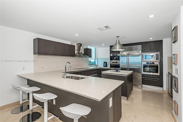 kitchen with built in appliances, sink, hanging light fixtures, a breakfast bar, and wall chimney exhaust hood