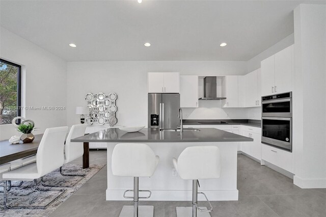 kitchen with a kitchen island with sink, sink, appliances with stainless steel finishes, wall chimney exhaust hood, and white cabinetry