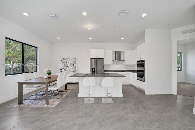 kitchen featuring a kitchen island with sink, stainless steel appliances, light tile floors, white cabinets, and wall chimney exhaust hood