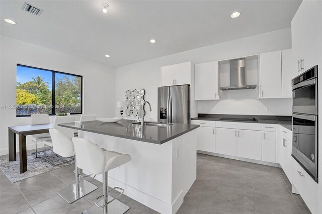 kitchen with wall chimney exhaust hood, stainless steel appliances, white cabinetry, and a kitchen island with sink