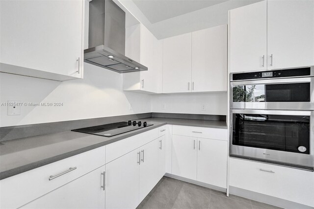 kitchen featuring stainless steel double oven, light tile floors, white cabinets, wall chimney range hood, and black electric stovetop