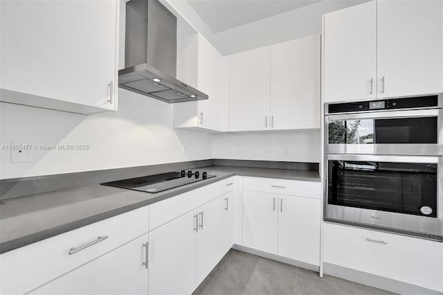 kitchen with black electric cooktop, dark countertops, white cabinetry, wall chimney range hood, and double oven