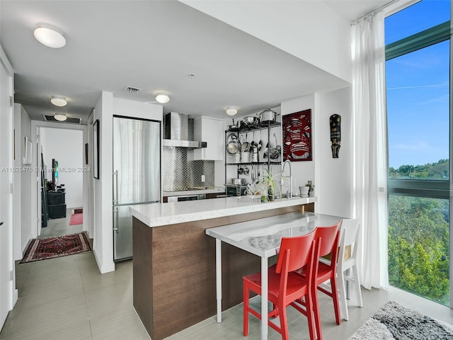 kitchen with kitchen peninsula, a breakfast bar area, tasteful backsplash, wall chimney range hood, and stainless steel fridge