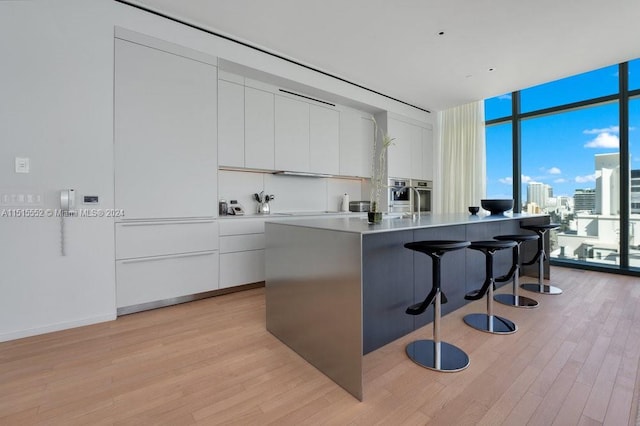 kitchen featuring light wood-type flooring, white cabinets, expansive windows, and an island with sink