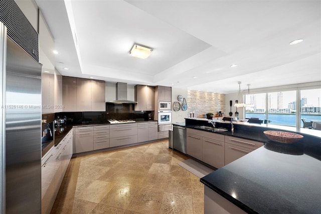 kitchen featuring gray cabinetry, backsplash, a tray ceiling, wall chimney range hood, and built in appliances