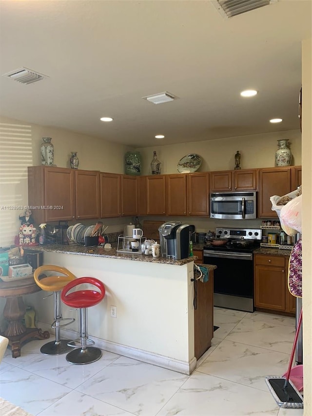 kitchen featuring kitchen peninsula, stainless steel appliances, light tile floors, dark stone countertops, and a breakfast bar area