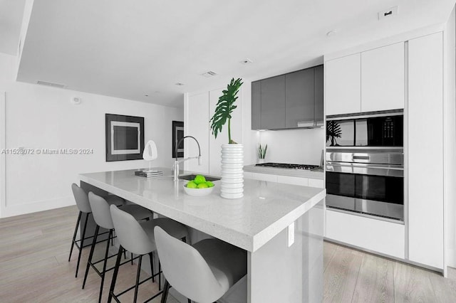 kitchen with gray cabinetry, a center island with sink, a kitchen bar, black microwave, and light wood-type flooring