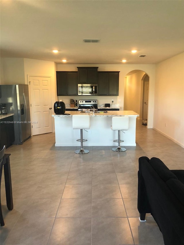 kitchen featuring a kitchen island with sink, light tile floors, appliances with stainless steel finishes, and a breakfast bar area