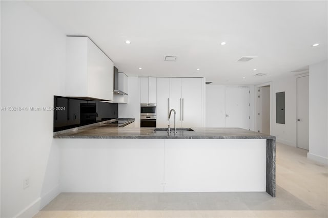 kitchen featuring stainless steel double oven, sink, dark stone countertops, white cabinets, and wall chimney range hood