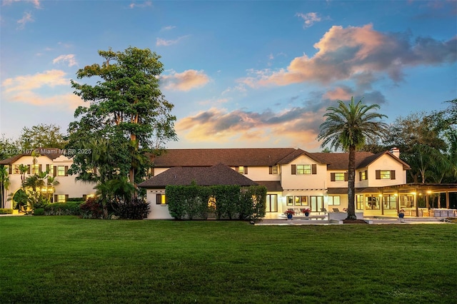 back house at dusk featuring a lawn and a patio