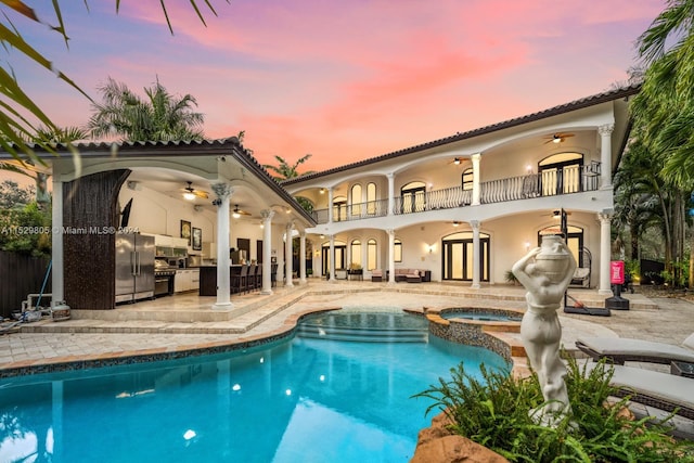 back house at dusk with a balcony, a swimming pool with hot tub, ceiling fan, and a patio