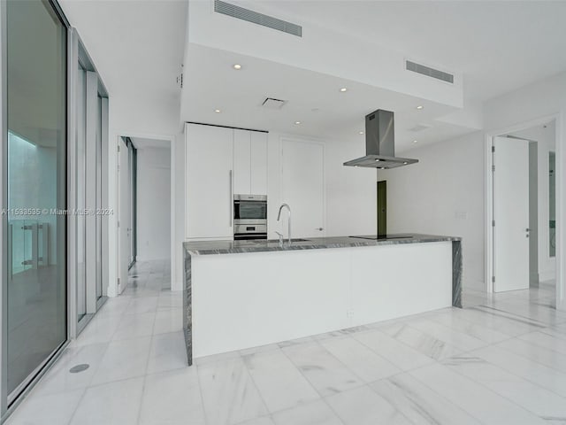 kitchen with double oven, dark stone counters, light tile flooring, white cabinetry, and island exhaust hood