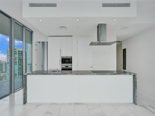 kitchen featuring island exhaust hood, white cabinets, double oven, light tile flooring, and stone countertops