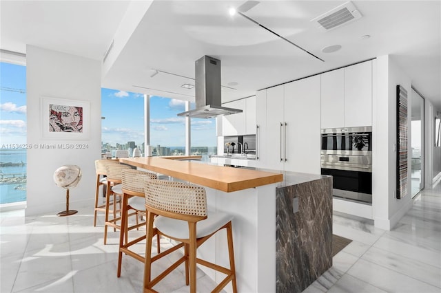 kitchen featuring a breakfast bar area, white cabinetry, island range hood, backsplash, and double oven