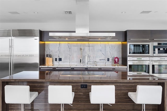 kitchen with built in appliances, sink, island exhaust hood, a breakfast bar, and tasteful backsplash