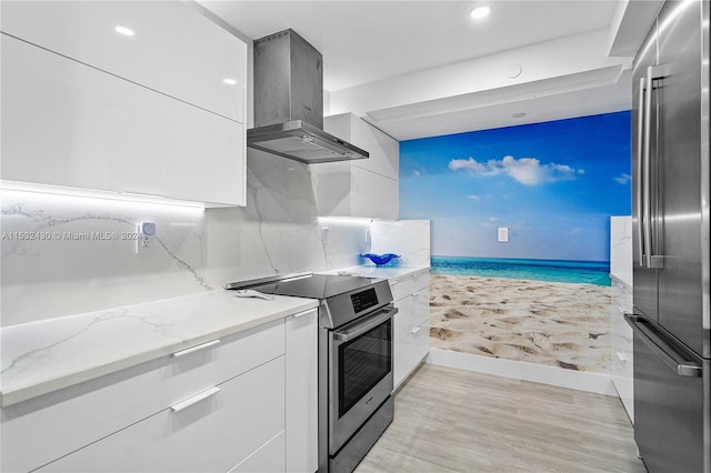 kitchen featuring white cabinetry, wall chimney range hood, light stone counters, appliances with stainless steel finishes, and light wood-type flooring