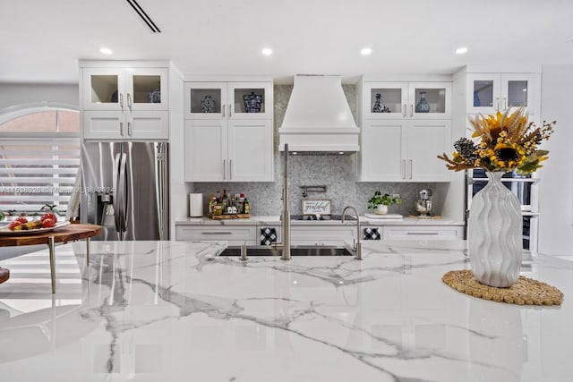 kitchen featuring custom exhaust hood, light stone countertops, white cabinets, backsplash, and stainless steel refrigerator with ice dispenser