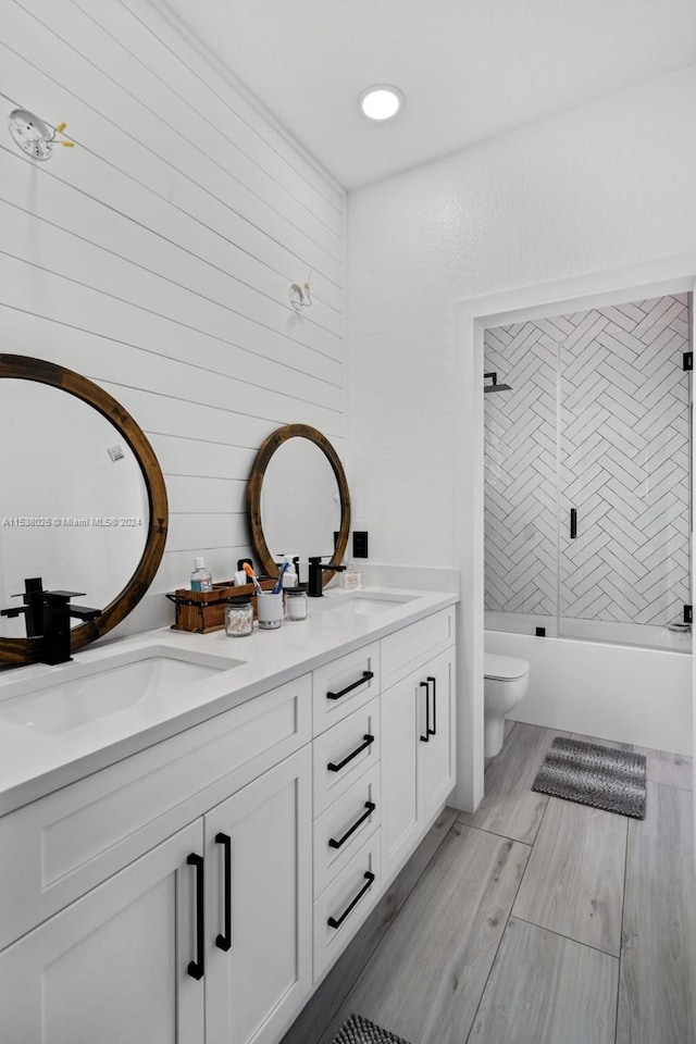 full bathroom with toilet, double sink, vanity with extensive cabinet space, bath / shower combo with glass door, and hardwood / wood-style flooring