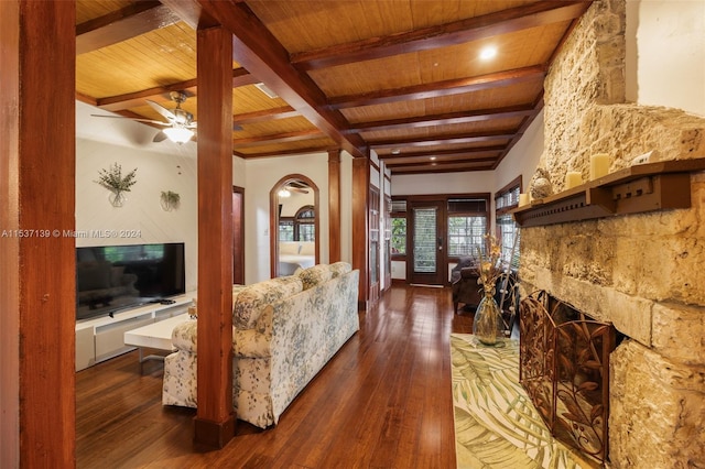 living room featuring a stone fireplace, wood ceiling, beamed ceiling, dark wood-type flooring, and ceiling fan